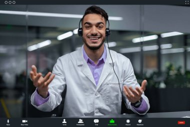 Beyaz önlüklü bir adam ofis ortamında görüntülü konuşma yaparken gülümsüyor ve jestler yapıyor. Meşgul görünüyor ve mesai saatleri içinde uzaktan biriyle iletişim kuruyor..