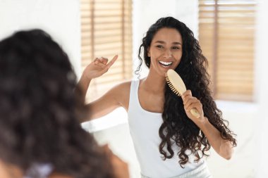 Uzun kıvırcık saçlı bir kadın aynanın önünde saçını tararken gülümsüyor. Güneş ışığı pencereden akar ve neşeli bir atmosfer yaratır..