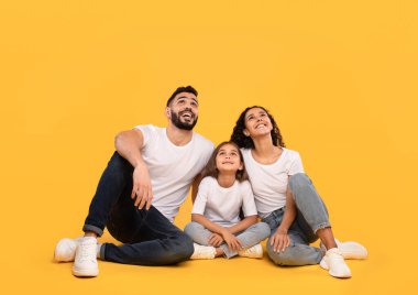 Üç kişilik bir aile yerde oturuyor, gülümsüyor ve heyecanla yukarı bakıyor. Neşeli ifadeleri bu neşeli anın tadını çıkarırlarken mutluluğu yansıtıyor..