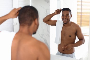 Parlak bir banyoda saçını taramak için tarak kullanan bir adam. Omuzlarının üzerinde bir havlu yatarken yansımasına gülümsüyor, sabah güneşinin tadını çıkarıyor..