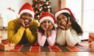 Aile üyeleri Noel için dekore edilmiş bir oturma odasında halıya uzanmış yatarken gülümserler. Kırmızı şapkalar takıyorlar ve etraflarını hediyelerle sarıyorlar, birlikte neşeyi paylaşıyorlar..