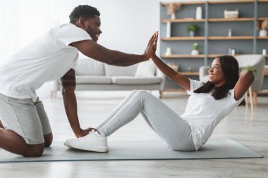 Evde karın kasları üzerinde çalışan mutlu, genç, siyah bir çift, kadın bacakları tutan, oturma odasında beşlik çakan bir adam. Spor, spor, karantina süresince egzersiz.