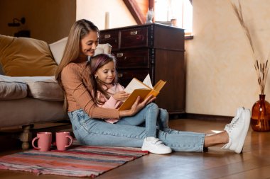 Bir anne ve kızı yerde oturup kitap okuyorlar. Yumuşak ışıklandırması ve rahat bir atmosferi olan sıcak, rahat bir oturma odasıyla çevrili..