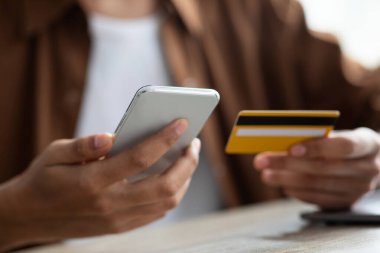 Bireyler bir masada oturuyorlar, bir elinde akıllı telefon, diğer elinde kredi kartı ile, online bir işlemi tamamlamak üzereler. Doğal ışık parıldıyor, rahat bir atmosfer yaratıyor..