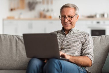 Son sınıf öğrencisi rahat bir koltukta oturuyor ve dizüstü bilgisayarını kullanırken gülümsüyor. Oturma odasının modern tasarımı ile sıcak bir atmosferi var, rahat bir ortam yaratıyor..