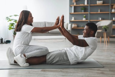 Gülümseyen siyah çift evde birlikte egzersiz yapıyor, karın kasları üzerinde çalışıyor. Neşeli Afrikalı Amerikalı kadın, erkeklerin bacaklarının üzerinde oturmuş karın ve karın kasları için egzersiz yaparken beşlikle tezahürat yapıyor.