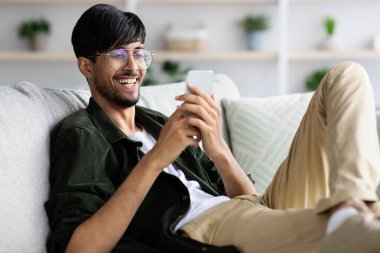 Genç bir adam rahat bir şekilde kanepede oturuyor, akıllı telefonuna bakarken gülümsüyor parlak bir oturma odasında bitkilerle ve yumuşak dekorla süslenmiş..
