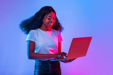 Kıvırcık saçlı genç bir kadın dizüstü bilgisayarla çalışırken eğleniyor. Renkli arka plan, gün boyunca modern bir çalışma alanı atmosferini yansıtan canlı bir dokunuş ekliyor..