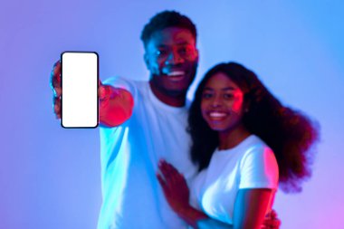 İki genç yetişkin birlikte poz veriyor, gülümsüyor ve önlerinde boş bir akıllı telefon tutuyor. Arka planda mavi ve pembe tonlarda parlak bir ışık var, canlı bir atmosfer yaratıyor..