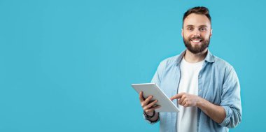 Genç bir adam elinde bir tablet tutarken güvenle ayakta duruyor. Sakalı var ve ceketiyle açık renk bir gömlek giyiyor. Arka plan parlak mavi bir renktir, neşeli atmosferi güçlendirir..