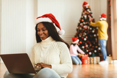 Bir kadın Noel şapkası takarken dizüstü bilgisayar kullanarak yerde oturuyor. Arkasında, aile üyeleri bir Noel ağacı süsler ve hediyeleri paketler..