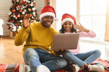 İki aile ferdi yerde dizüstü bilgisayarla oturup ekrana el sallıyor. Noel şapkası takıyorlar ve Noel süsleri ve Noel ağacıyla çevrili bir video görüşmesinden zevk alıyorlar..