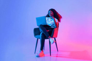 Genç bir kadın, dizüstü bilgisayarıyla meşgul bir şekilde bir sandalyede rahatça oturuyor. Arka planda canlı mavi ve pembe renkler yer alıyor. Odaklanmış ve rahat görünüyor, davetkar bir atmosfer yaratıyor..