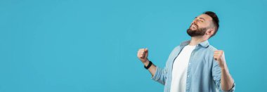 Yumruklarını zaferle kaldırmış bir adam büyük bir gülümseme gösterir. Parlak mavi arka plan mutluluk duygusunu arttırır. Bu an saf neşe ve kutlamayı yakalar..