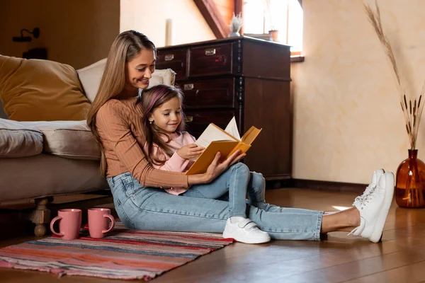 Bir anne ve kızı yerde oturup kitap okuyorlar. Yumuşak ışıklandırması ve rahat bir atmosferi olan sıcak, rahat bir oturma odasıyla çevrili..
