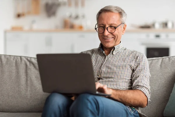 Son sınıf öğrencisi rahat bir koltukta oturuyor ve dizüstü bilgisayarını kullanırken gülümsüyor. Oturma odasının modern tasarımı ile sıcak bir atmosferi var, rahat bir ortam yaratıyor..