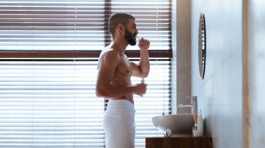 Bir adam şık bir banyoda duruyor, havluyla dişlerini fırçalıyor. Güneş ışığı panjurların arasından parlıyor ve uzayda sıcak bir atmosfer yaratıyor..