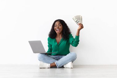 Neşeli bir kadın önünde dizüstü bilgisayarla yerde oturuyor. Elinde birkaç dolarlık banknotlar tutarken büyük bir gülümseme ile finansal başarısını gösteriyor..