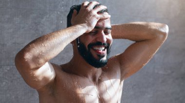 Sıcak bir duşun altında duran bir adam. Vücudundan aşağı çağlayan bir su. O, ellerini saçlarında gezdirirken neşeyle gülümser. Güneş ışığı filtreler ve rahatlatıcı bir atmosfer yaratır..