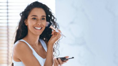 Kıvırcık saçlı bir kadın aydınlık bir odada makyaj yapıyor. Yanaklarına allık sürmek için fırça kullanır, kendine güvenerek gülümser, güzellik rutininin tadını çıkarır ve kendini güçlü hisseder..