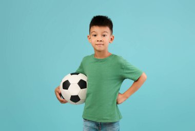Kısa siyah saçlı bir çocuk, sol tarafında siyah ve beyaz bir futbol topu tutan, kendinden emin bir şekilde duruyor. Açık mavi arka planda yeşil gömlek ve kot pantolon giyiyor..