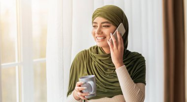 Yeşil eşarp takan genç bir kadın telefonda konuşurken gülümsüyor. Bir kupa kahve tutuyor ve pencerenin yanında duruyor, içeri güneş ışığı giriyor..