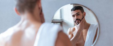 Bir adam yüzüne hafifçe dokunarak yansımasını inceler. Beline sarılı bir havlu var ve duş aldıktan sonra parlak, çağdaş bir banyoda duruyor..