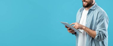 Sakallı bir adam tabletle mutlu bir şekilde etkileşim içindedir. Açık renkli bir gömlek giyer ve mavi bir arka plana yaslanır. Dijital deneyiminin tadını çıkarır..