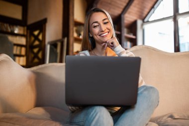 Genç bir kadın, dizüstü bilgisayarını kullanırken rahat bir şekilde kanepede oturur ve gülümser. Oturma odasının sıcak, davetkar atmosferi ahşap aksanlar ve doğal ışık barındırır..