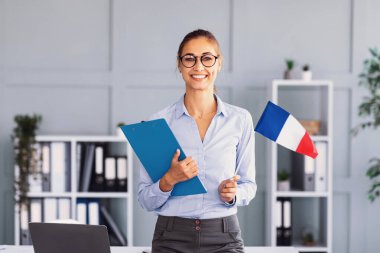 Kendine güvenen bir kadın parlak bir ofiste elinde mavi bir dosya ve kalemle kameraya gülümsüyor. Küçük bir Fransız bayrağı sergileniyor ve profesyonel atmosfere ekleniyor.