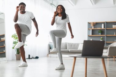 Ev antrenmanı. Gülümseyen siyah adam ve kadın koca oturma odasında birlikte antrenman yapıyor, dirsek egzersizi yapıyor. Antrenörle sanal derste ayağa kalkan ve kaldıran mutlu çift.