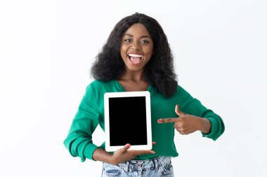 Kıvırcık saçlı neşeli bir kadın boş bir tablet tutuyor ve onu işaret ediyor. Beyaz bir arka planın önünde duran yeşil bir gömlek ve kot pantolon giyiyor..