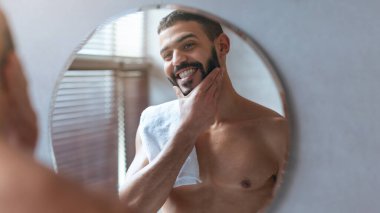 Bir adam yuvarlak aynanın önünde duruyor, sakalına dokunurken gülümsüyor. Üstü çıplak ve boynunda bir havlu var. Güneş ışığı parlak banyoyu dolduruyor..