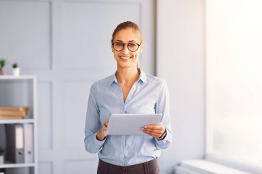 Gözlüklü, kendine güvenen bir kadın iyi aydınlatılmış bir ofiste elinde tablet tutarken gülümsüyor. Gösterişli alanda hafif duvarlar ve organize raflar var, profesyonel bir atmosfer yaratıyor..