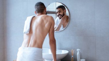 Yuvarlak aynanın önünde duran bir adam havluyla yansımasını inceliyor. Banyo ayarları parlak ve modern, temiz lavabo ve tuvalet malzemelerini gösteriyor..