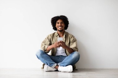 Genç siyah bir adam beyaz bir duvarın yanında yerde rahat bir şekilde oturuyor, akıllı telefonunu kullanırken gülümsüyor. Uygulamaları gezerken ya da arkadaşlarına mesaj atarken mutlu görünüyor. Rahatlatıcı bir atmosfer yaratır..