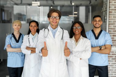 Çok ırklı doktor ekipleri üniformalı erkekler ve kadınlar sabah erken saatlerde klinikte ya da tıbbi konferansta birbirlerine poz veriyorlar, kameraya neşeli bir şekilde gülümsüyorlar, erkek hekim baş parmağını kaldırıyor.