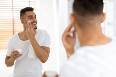 Bir adam, yansımasına gülümserken parmaklarıyla yüzüne nemlendirici sürer. Banyo aydınlık ve havadar. Bakım için temiz bir atmosfer yaratıyor..