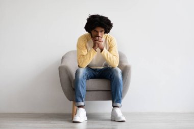 Genç bir Afrikalı Amerikalı evdeki koltukta sessizce oturur, derin düşüncelere dalmış. Yüzündeki ifade, zihinsel sağlığıyla ilgili kişisel mücadelelerle uğraşırken üzüntüyü ve tefekküre neden olur..