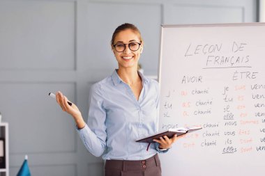 Bir öğretmen beyaz tahtanın önünde duruyor, öğrencileri Fransızca dersiyle meşgul ediyor. Fransızca öğrenmek için anahtar sözcük dağarcığını sunarken kendine güvenen bir not defteri ve jestler tutuyor..