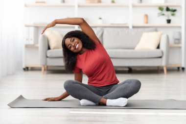 Genç bir kadın, oturma odasındaki minderde yoga yaparken vücudunu esnetiyor. Sıcacık bir ortamda sağlığın ve farkındalığın tadını çıkarıyor..