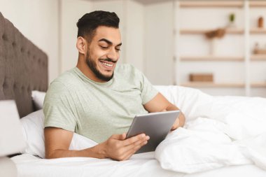 Bir adam tabletle etkileşime girerken rahatça yatakta oturup gülümsüyor. Yumuşak yatak örtüsü onu doğal ışıkla dolu, parlak, rahat bir yatak odasına sarmış..