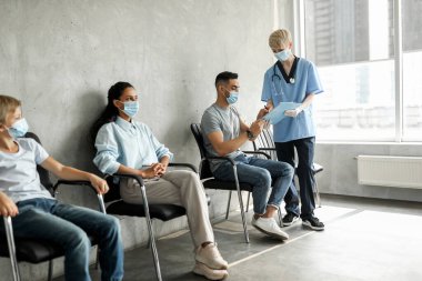 Orta Doğulu genç hasta aşılanmadan önce kağıtları dolduruyor. Kadın doktor, yetişkin ve okul çağındaki insanlara yardım ediyor. Koronavirüse karşı aşı bekliyorlar.