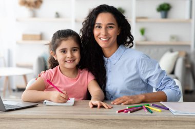 Bir anne, kızı pembe bir kalem kullanarak deftere çizerken gururla gülümser. Parlak bir yaşam alanındaki renkli kırtasiyelerle çevrili ahşap bir masada oturuyorlar..