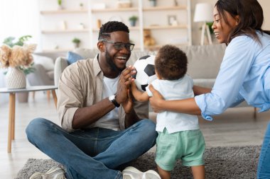 Neşeli bir siyahi aile evde oyun oynuyor. Ebeveynler bebeklerinin elinde bir futbol topu tutarken gülümserler, yerde otururken. Bu anda aile bağları ve neşeli ebeveynlik sergileniyor..