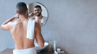 Bir adam şık bir banyoda, belinde havluyla dikilip aynadaki yansımasına gülümsüyor. Kendine güveni ve özgüveni var..