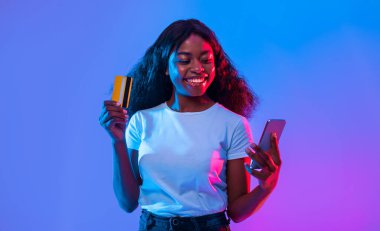 Bir kadın canlı bir ortamda, elinde kredi kartı tutarken telefonuna sevinçle bakıyor. Renkli ışıklandırma modern alışveriş alışkanlıklarını yansıtan canlı bir atmosfer yaratıyor..