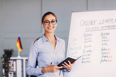 Almanca notlarla dolu bir tahtanın yanında bir öğretmen duruyor. Bir defter tutuyor ve kulaklık kullanarak gülümsüyor..