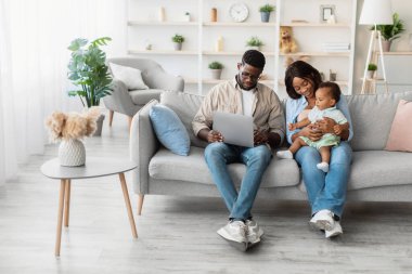 Genç siyahi ebeveynler kanepede dizüstü bilgisayar kullanırken küçük oğullarıyla mutlu bir şekilde etkileşime geçiyorlar. Eğlenceli bir eğitim oyunuyla meşguller. Parlak oturma odalarında dinleniyorlar..