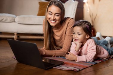 A mother and her young daughter are lying on a colorful rug, engaged with a laptop. They share smiles and laughter, creating a warm and joyful atmosphere in their cozy living room.